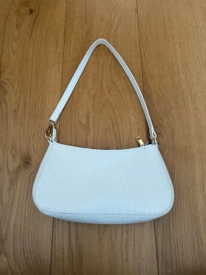 Vit axelremsväska i crocopräglat material - Säljer en stilren vit handväska med axelrem och crocopräglat mönster. Väskan har guldfärgade metalldetaljer och dragkedja upptill. Insidan är svartfodrad och modellen är kompakt och smidig, perfekt för det viktigaste. Materialet känns som konstläder.