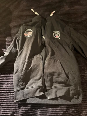 Svart zip-hoodie från Vinson Polo Club - Svart hoodie med dragkedja från Vinson Polo Club. Har broderade loggor på bröstet, två stora fickor fram och vita snören i huvan. Perfekt för en avslappnad och sportig stil. Mjuk bomullskänsla och ribbade muddar vid ärmslut och nederkant.