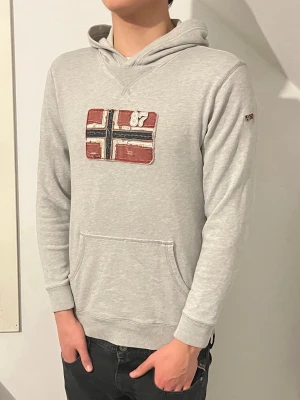 Ljusgrå hoodie från Napapijri - En ljusgrå hoodie från Napapijri. Inga defekter alls och har aldrig varit använd. Storleken på denna hoodien är en S/M. Modellen i bilden är 188cm lång och väger 74kg. 