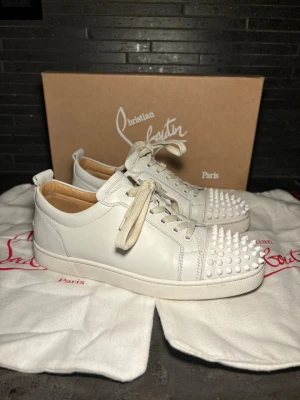 Vita Christian Louboutin sneakers  - Säljer ett par vita sneakers från Christian Louboutin med coola vita nitar på tån. Skorna är låga, har vita skosnören och är tillverkade i läder. Perfekta för dig som vill sticka ut med en lyxig och edgy stil. Storlek 40 sitter 41 allt og 