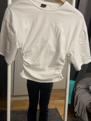Vit croppad topp från Gina - Snygg vit croppad topp från Gina med korta ärmar och lätt rynkade sidor. Enkel och fräsch design i mjuk bomull, perfekt till jeans eller kjol. Rund halsringning och relaxed fit för en chill vibe.