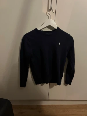 Mörkblå tröja från Ralph Lauren - Snygg mörkblå tröja från Ralph Lauren med klassisk rund halsringning och den ikoniska broderade loggan på bröstet. Tröjan har långa ärmar och är tillverkad i ett mjukt, stickat material som känns skönt mot huden. M (barn) storlek 