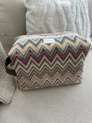 Mönstrad necessär från Emelo - Necessär i tyg från Emelo med färgglatt zickzackmönster i beige, rosa, vinrött och svart. Rektangulär form med vit dragkedja och brunt handtag i sidan. Perfekt för smink eller småprylar. 