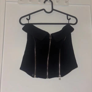 Svart korsett med dragkedjor - Trendig svart korsettopp med bred krage och tre silvriga dragkedjor framtill. Snygg croppad modell som ger edgy vibe, perfekt att styla med höga jeans eller kjol. Materialet känns stadigt och håller formen.