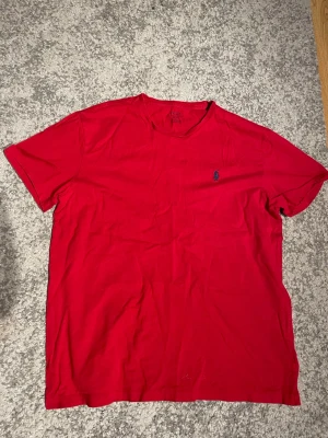 Röd t-shirt från Ralph Lauren - Röd t-shirt från Ralph Lauren med klassisk passform och liten blå broderad logga på bröstet. T-shirten har rund halsringning och är tillverkad i mjuk bomull. Perfekt för en avslappnad och stilren look.