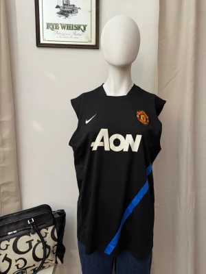 Manchester United x Nike svart träningslinne - Snyggt svart träningslinne från Nike med Manchester Uniteds klubbmärke på bröstet och AON-logga i vitt. Linnet har en blå diagonal detalj och är ärmlöst med rund hals. Perfekt för fotbollsträning eller gymmet.