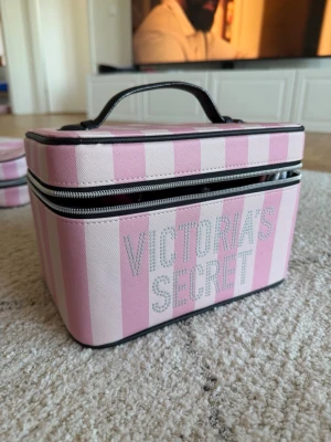 Victoria's Secret rosa necessär - Snygg necessär från Victoria's Secret i rosa och ljusrosa ränder med glittrande logga framtill. Väskan är boxformad med svart dragkedja och handtag upptill. Perfekt för smink eller småprylar, tillverkad i tåligt tygmaterial.
