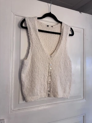Ljus stickad väst/topp med knappar H&M - Snygg ljus stickad väst/topp med v-ringning och pärlemorskimrande knappar framtill från H&M. Toppen är ärmlös och har en croppad passform med ribbad nederkant. Perfekt att styla över en skjorta eller t-shirt för en trendig look.
