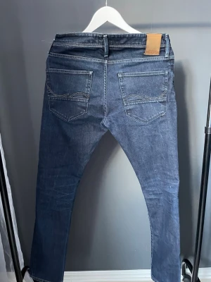Mörkblå jeans från Jack & Jones - Säljer ett par mörkblå raka jeans från Diesel med klassiska fem fickor och snygga slitningar framtill. Jeansen har läderpatch med logga bak i midjan och diskreta kontrastsömmar. Perfekta för en avslappnad och trendig look.