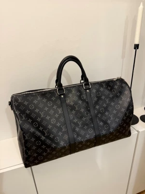 Louis Vuitton Keepall 55 svart monogram  - Snygg och rymlig resväska från Louis Vuitton i svart med klassiskt monogrammönster i grått. Väskan har två robusta handtag, silverfärgade metalldetaljer och är tillverkad i slitstarkt canvasmaterial. Perfekt för weekendresor och äventyr.