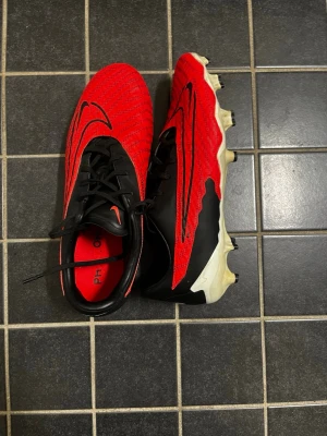 Nike Phantom röda fotbollsskor - Säljer ett par Nike Phantom Gx fotbollsskor i rött och svart med vita detaljer. Skorna har snörning, syntetiskt material och mönstrad ovandel för bättre bollkontroll. Sulan har fasta dobbar för spel på naturgräs. Snygg och modern design med stora Nike-loggor.