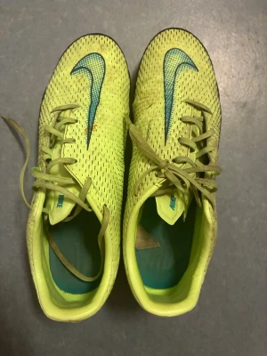 Gula Nike Mercurial fotbollsskor - Säljer ett par gula Nike Mercurial fotbollsskor med blå Nike-logga och snörning. Skorna har dobbar för spel på gräs och är tillverkade i syntetmaterial med ventilerande mesh. Perfekta för dig som vill sticka ut på planen!