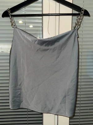 Silver kortkjol med kedjedetaljer - Snygg silverfärgad kortkjol med glansigt tyg och kedjor som axelband. Kjolen har en enkel, rak passform och ger en cool statement-look. Perfekt för dig som vill sticka ut med en unik detalj i outfiten.