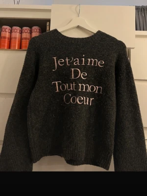 Mörkgrå stickad tröja Gina Tricot - Mysig mörkgrå stickad tröja från Gina Tricot med texten 'Je t’aime de tout mon coeur' broderad framtill i ljusrosa. Tröjan har rund halsringning, långa ärmar och ribbade muddar. Perfekt för dig som gillar franska vibes och sköna plagg.