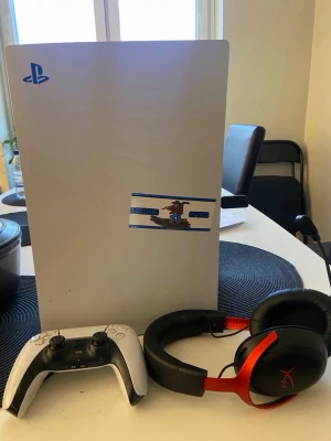 PlayStation 5 med kontroll och headset - Säljer en PlayStation 5 konsol i vit och svart med blå PlayStation-logga och en unik klisterdekal på framsidan (tas bort). Medföljer en vit och svart DualSense-kontroll samt ett HyperX gamingheadset i svart och rött. Perfekt för gaming och streaming.