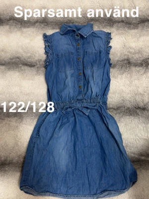 Blå jeansklänning med volangärmar - Snygg blå jeansklänning med volangdetaljer vid ärmarna, knappar framtill och två bröstfickor. Klänningen har knyt i midjan för en skön passform och är ärmlös. Perfekt för en avslappnad och trendig look.🌸 Sparsamt använd. Storlek 122/128 