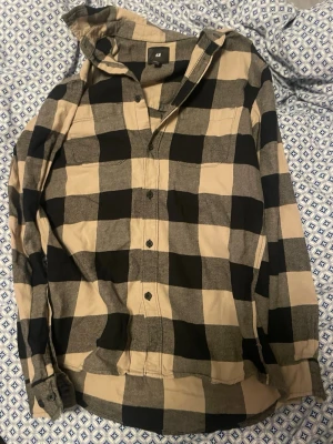 Rutig skjorta från H&M i beige och svart - Säljer en snygg rutig skjorta från H&M i beige och svart. Skjortan har klassisk krage, knappar framtill och långa ärmar med knapp vid ärmslut. Perfekt för en avslappnad och trendig look.