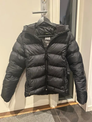 Svart pufferjacka från Helly Hansen - Säljer en super snygg jacka från Helly Hansen med huva och dragkedja framtill. Jackan har två sidofickor med dragkedja och breda, vadderade paneler för extra värme. Perfekt för kalla dagar och riktigt skön passform. Säljer pågrund av att den blivit för liten och såklart är jackan tvättad
