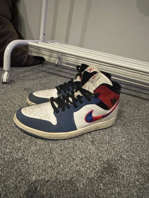 Nike Air Jordan 1 Mid blå/röd/vit - Snygga Nike Air Jordan 1 Mid sneakers i vitt, blått och rött med svarta skosnören. Klassisk Jordan-logga på plösen och röd Swoosh på sidan. Ovandel i läder och textil, perforerad tå för extra ventilation. Perfekta för dig som gillar streetwear och basketstil.