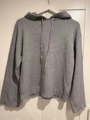 Grå stickad hoodie med huva  - En basic grå hoodie med huva. Hoodien har en avslappnad passform, långa ärmar och ribbade muddar vid ärmslut och nederkant. Perfekt för en chill och enkel stil. Materialet känns mjukt och bekvämt.