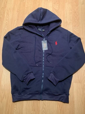Marinblå hoodie från Polo Ralph Lauren - Mörkblå hoodie med dragkedja från Polo Ralph Lauren. Klassisk design med huva och fickor framtill, samt röd broderad logga på bröstet. Tillverkad i mjukt bomullsmaterial för en bekväm känsla. Perfekt för en avslappnad och stilren look.