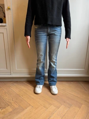 Blå bootcut jeans från Diesel - Säljer ett par klassiska blå bootcut jeans från Diesel med snygg tvätt och lätt slitning. Jeansen har fem fickor, normal midja och dragkedja med knapp. Perfekta för en avslappnad och trendig look. Storlek 27 Klassiska Diesel jeans i mellanblå tvätt med stilren bakficka och diskret röd logodetalj. Rak modell med lätt bootcut som ger fin silhuett och balans. Mjuk denim med stretch som formar sig efter kroppen och sitter bekvämt hela dagen. Modellen är 160 cm lång och väger 50 kg.