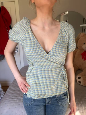 Gulblå rutig omlottblus - Söt omlottblus med puffiga korta ärmar i gulblått rutigt mönster. Djup v-ringning framtill och knytband i midjan för en feminin siluett. Tunt, luftigt material som passar perfekt till jeans eller kjol.