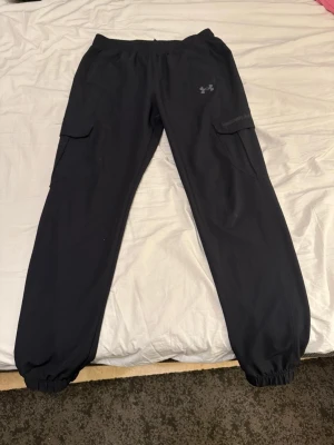 Svarta mjukisbyxor från Under Armour - Mörka mjukisbyxor från Under Armour med elastisk midja och muddar vid bensluten. Byxorna har två stora cargofickor på sidorna och en diskret logga på vänster lår. Perfekta för en chill och sportig look.