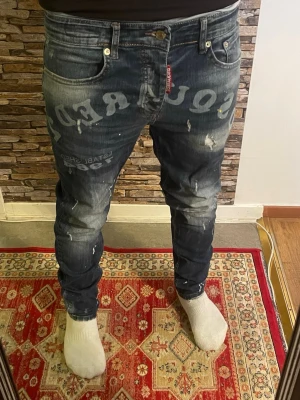 Mörkblå Dsquared2 jeans med tryck - Säljer ett par mörkblå jeans från Dsquared2 med stora vita tryck och slitningar. Jeansen har skinny fit, klassiska fem fickor och detaljer som färgstänk och distressed look. Perfekta för dig som gillar streetstyle och vill sticka ut.