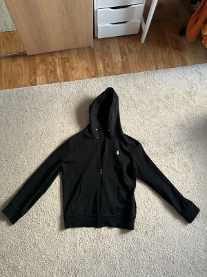 Svart hoodie från Polo Ralph Lauren - Säljer en svart hoodie från Polo Ralph Lauren med dragkedja framtill och den klassiska vita loggan broderad på bröstet. Hoodien har huva med dragsko och ribbade muddar. Perfekt för en chill och stilren look. R€P är den men av väldigt bra kvalitet