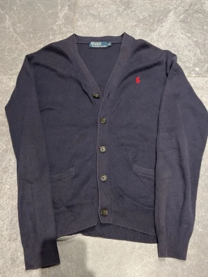 Mörkblå cardigan från Polo Ralph Lauren - Mörkblå cardigan med knappar från Polo Ralph Lauren. Klassisk v-ringning, två fickor framtill och röd broderad logga på bröstet. Pris går att diskutera vid snabb affär 