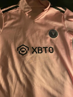 Inter Miami Messi matchtröja rosa - Snygg rosa matchtröja från Inter Miami med Messi och nummer 10 på ryggen. Tröjan har svarta detaljer, klubbmärke på bröstet och sponsorlogga framtill. Tillverkad i lätt och ventilerande material, perfekt för fotboll eller träning.