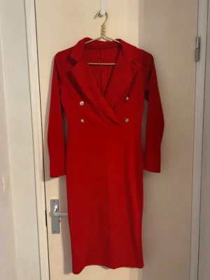 Röd långärmad klänning med knappar - Stilren röd klänning med lång ärm och markerad krage. Klänningen har dubbelknäppning med dekorativa silvriga knappar framtill och en figurnära passform. Perfekt för dig som vill sticka ut med en klassisk look.