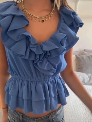 Blå volangblus från H&M - Supersöt blå blus från H&M med volanger längs urringningen och nedtill. Ärmlös modell med v-ringad hals och resår i midjan som ger en feminin siluett. Perfekt för dig som vill ha en trendig och lekfull topp till sommaren.