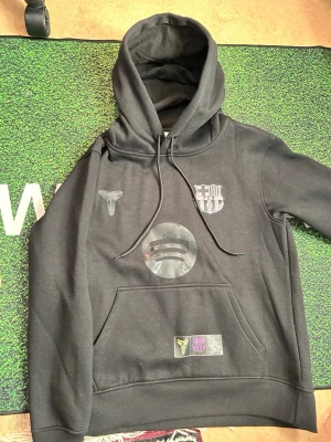 Svart FC Barcelona x Kobe Bryant hoodie - Snygg svart hoodie med FC Barcelona-logga i svartvitt och Kobe Bryants logga på bröstet. 