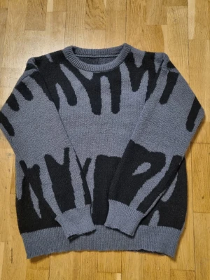 Grå stickad tröja med svart mönster - Säljer en grå stickad tröja med abstrakt svart mönster. Tröjan har rund halsringning, ribbade muddar och avslut. Perfekt för dig som gillar unika plagg med grafisk design. Materialet känns mjukt och passar bra till lager på lager.
