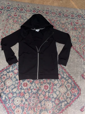 Svart hoodie med dragkedja från BOSS - Svart hoodie från BOSS med dragkedja framtill och diskret logga på bröstet. Klassisk modell med huva och långa ärmar. Perfekt för en chill och stilren look. Materialet känns mjukt och bekvämt, troligen bomullsblandning.