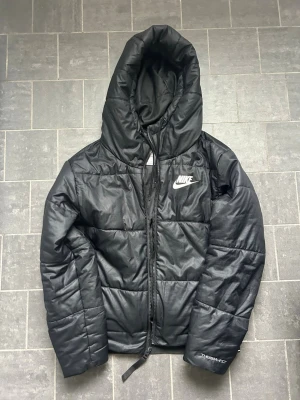 Svart pufferjacka från Nike - (RUNT 700 KR NYPRIS) Säljer en snygg svart pufferjacka från Nike med stor huva och dragkedja framtill (knappt använd). Jackan har Nike-logga på bröstet och Therma-FIT-märkning på ärmen. Tillverkad i ett glansigt syntetmaterial som håller värmen under kalla dagar. Perfekt för dig som vill ha en snygg jacka under hela året. Ligger runt 700 kr nya, är du intresserad så är det bara att skriva så löser vi den till dig direkt 🍾✈️