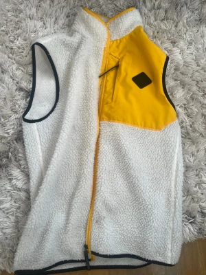 Vit/gul J. Lindeberg  - Säljer en riktigt snygg vit Lindeberg fleeceväst för jag behöver pengar. med gul bröstficka och svart kantband. Västen har dragkedja framtill och en cool detalj med svart logotyp på bröstet. Perfekt för lager-på-lager och chill stil. Materialet är mjuk teddyfleece och nylon på fickan. Den är knappt andvänd. 