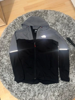 The North Face svart/grå vindjacka - Snygg vindjacka från The North Face i svart och grått med hel dragkedja och huva. Jackan har reflekterande detaljer på bröstet och ärmarna samt justerbar huva med röda snören. Perfekt för blåsiga dagar och streetwear-stil.sälj inte längre 