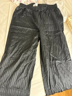 Svarta pyjamasbyxor med vita detaljer - Snygga svarta pyjamasbyxor med subtila ränder och vita passpoaldetaljer vid bensluten. Byxorna har resår i midjan för extra komfort och är tillverkade i ett mjukt, glansigt material som känns lyxigt mot huden. Perfekta för mysiga hemmakvällar.