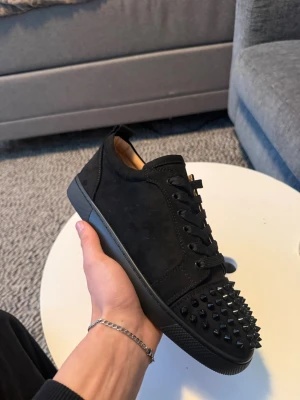 christian louboutin junior spiked - Snygga svarta sneakers i mocka från Christian Louboutin med svarta nitar på tån och svarta skosnören. Låg modell med klassisk siluett och exklusiv känsla. Insidan är i ljust läder och yttersulan är svart. Perfekta för dig som gillar edgy och lyxig stil.  Helt nya skickar de idag 