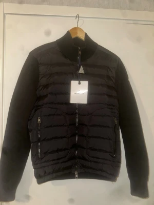 Svart cardiganjacka från Moncler - Helt ny Snygg svart cardiganjacka från Moncler med quiltad front och stickade ärmar. Jackan har hög krage, två dragkedjeförsedda fickor och Moncler-logga på ärmen. Perfekt för dig som vill ha en stilren och exklusiv look. Påse ingår.