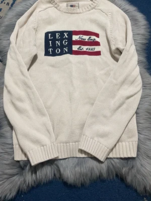 Vit stickad tröja från Lexington - Vit stickad tröja från Lexington med amerikanskinspirerad flaggdetalj och text på bröstet. Tröjan har rund halsringning, ribbstickade muddar och nederkant. Perfekt för dig som gillar klassisk och avslappnad stil. Tröjans ny pris är 1000 men säljs billigt eftersom den är liten för mig
