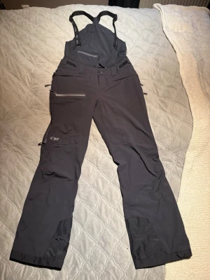 Svarta skalbyxor med hängslen OR - Svarta skalbyxor från Outdoor Research med hängslen och hög midja. Byxorna har flera dragkedjeförsedda fickor, förstärkta benslut och justerbara detaljer i midjan. Perfekta för vinterns äventyr och skidåkning. Modellen heter Skyward. Bib. Två små små hål nere vid ankeln på vänstra benet.