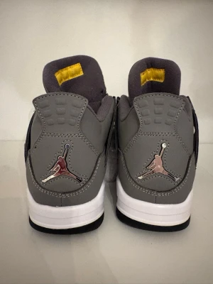 Gråa Air Jordan 4 Retro sneakers - Säljer ett par gråa Air Jordan 4 Retro sneakers med metallic Jumpman-logga på hälen och gula detaljer på plösen. Skorna har vit mellansula och svart yttersula. Tillverkade i läder och textil för en klassisk och clean look.   INTE ÄKTA