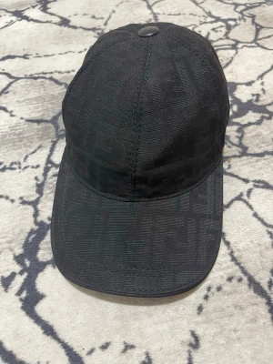 Svart keps från Fendi med FF-mönster - Snygg svart keps från Fendi med diskret FF-monogram i tyget. Kepsen har böjd skärm, justerbart spänne bak och är tillverkad i 100% bomull. Perfekt accessoar för en trendig och exklusiv look.