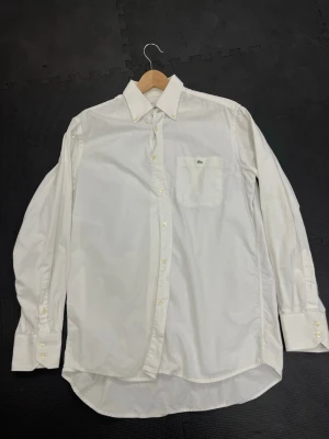  Lacoste Skjorta  - Klassisk vit skjorta från Lacoste med lång ärm, bröstficka och den ikoniska krokodilloggan. Skjortan har knappar framtill och vid ärmsluten, samt en tidlös krage. Perfekt för en clean och stilren look.
