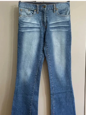 Blå bootcut jeans med ränder - Snygga blå bootcut jeans med tunna vita vertikala ränder och lätt slitning framtill. Klassisk femficksmodell med knapp och dragkedja. Jeansen har en trendig utsvängd passform och är tillverkade i denim.