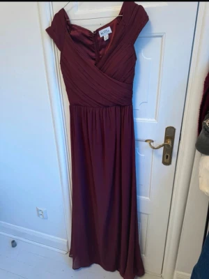 Vinröd långklänning från NLY Eve - Elegant vinröd långklänning från NLY Eve med omlottliv och korta ärmar. Klänningen har v-ringning och draperade detaljer upptill, samt en mjukt fallande kjol. Perfekt för speciella tillfällen.